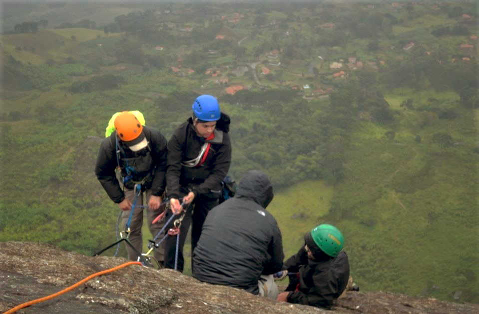 Fotografia colorida. Quatro escaladores parados no meio de uma rocha rugosa amarronzada. Os dois escaladores da esquerda estão em pé, de frente para a câmera, amarrados por cordas presas a sistemas de parada montados na rocha. O primeiro da esquerda usa capacete laranjado sobre boné, jaqueta preta e calça bege. O segundo usa capacete azul, jaqueta e calças pretos. Os dois escaladores da direita, estão sentados. O primeiro, de costas para a câmera, usa jaqueta preta com capuz cobrindo parte da cabeça. O segundo, pouco mais abaixo, usa capacete verde e jaqueta preta. Lá em baixo uma área verdejante com vegetações baixas, alguns amontoados de árvores e casas rodeando uma estrada.