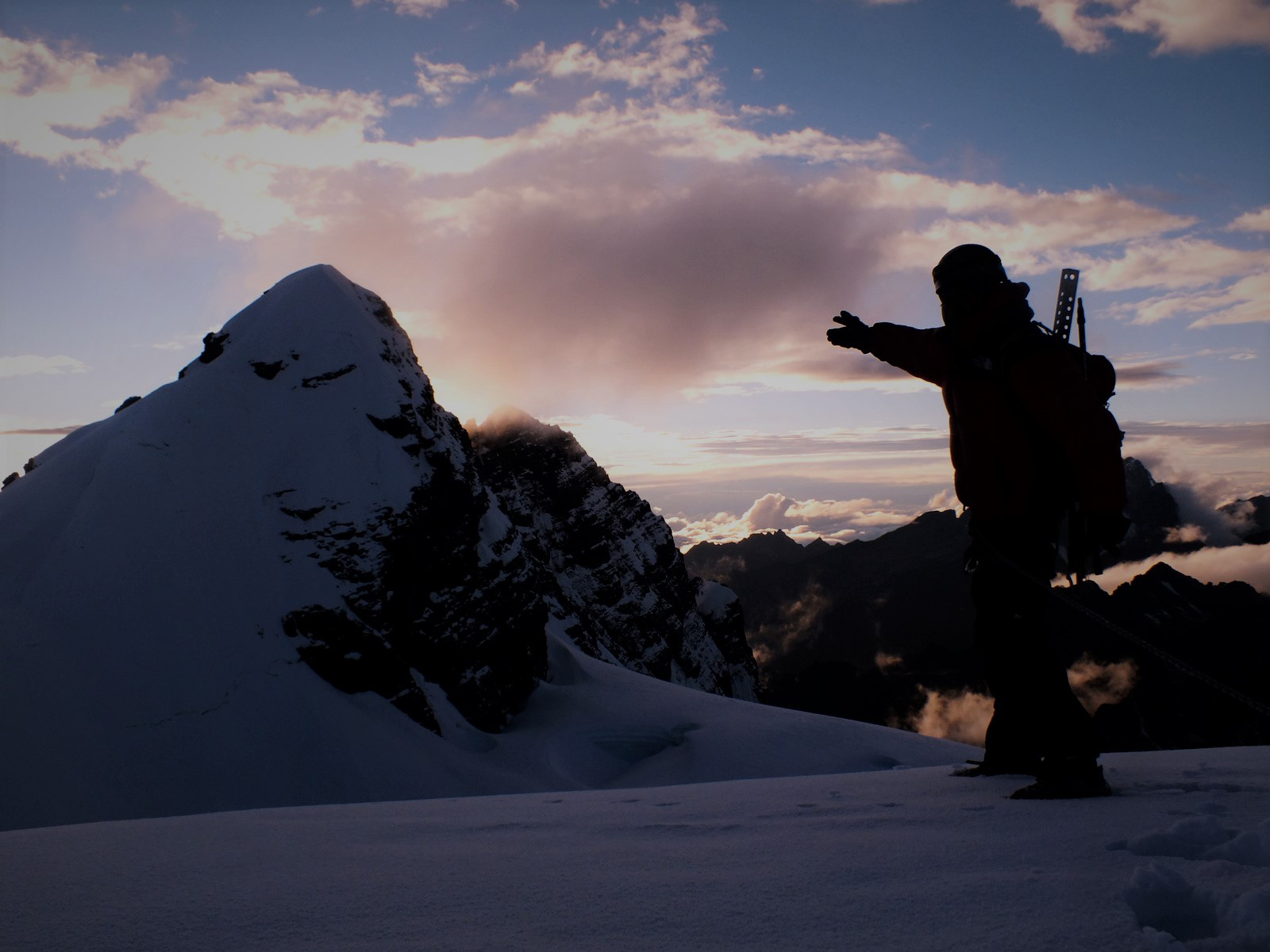 Fotografia colorida. Silhueta de um escalador apontando um pico montanhoso coberto de neve. Pela contraluz, o contorno da mochila com equipamentos que ele carrega nas costas. Ele está em pé sobre a neve e distante dele, do lado esquerdo da foto, o pico que sobressai de outras formações rochosas mais além por entre algumas nuvens. A luminosidade do sol é bloqueada pelo pico e por algumas nuvens mais altas.