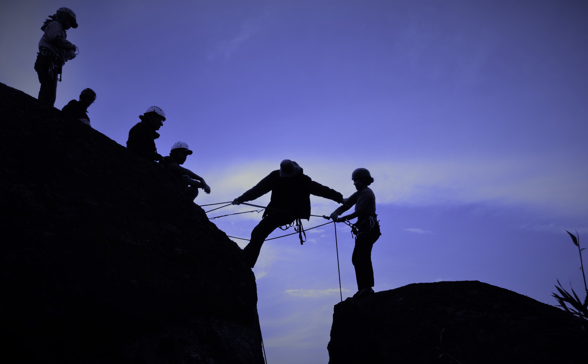 Fotografia colorida. Sobre céu azul, silhueta de um grupo de escaladores em duas pedras separadas por um vão. Preso a algumas cordas, um escalador se inclina estendendo um dos braços na direção da escaladora que está sozinha na pedra a direita da foto. Ela segura a corda amarrada a sua cadeirinha. Quatro escaladores sobre a pedra da esquerda observam os outros dois escaladores.